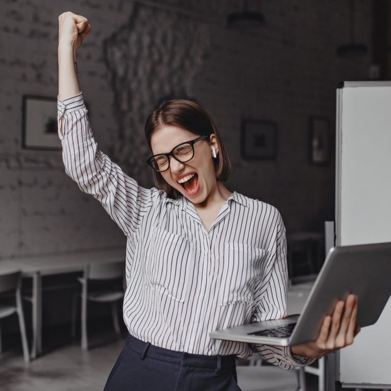 Business woman with laptop in hand is happy with success. Portrait of woman in glasses and striped blouse enthusiastically screaming and making winning gesture